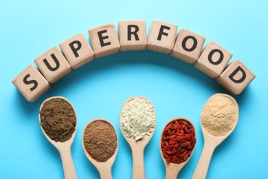 Photo of Different superfoods in spoons and word made of wooden cubes on light blue background, flat lay