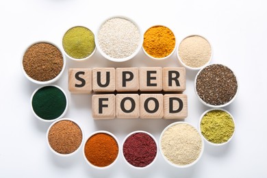 Photo of Different superfood powders in bowls and words made of wooden cubes on white background, flat lay