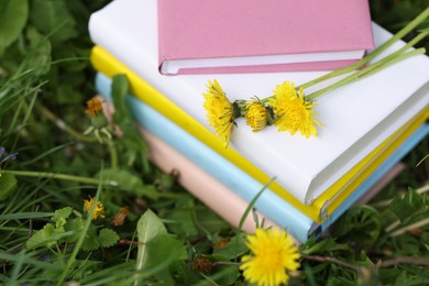 Photo of Stacked books with dandelion flowers on green grass outdoors, closeup