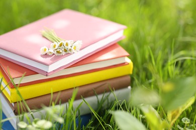 Photo of Stacked books with chamomile flowers on green grass outdoors, closeup