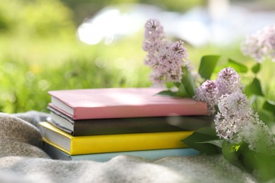 Photo of Stacked books, lilac flowers and blanket on green grass outdoors, closeup