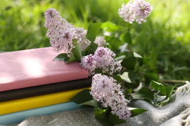 Photo of Stacked books, lilac flowers and blanket on green grass outdoors, closeup