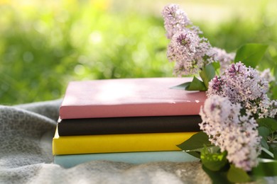 Photo of Stacked books, lilac flowers and blanket on green grass outdoors, closeup