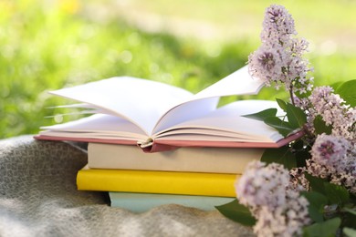 Photo of Stacked books, lilac flowers and blanket on green grass outdoors, closeup