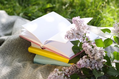 Photo of Stacked books, lilac flowers and blanket on green grass outdoors, closeup