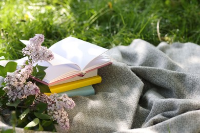 Photo of Stacked books, lilac flowers and blanket on green grass outdoors