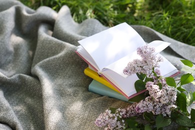 Photo of Stacked books, lilac flowers and blanket on green grass outdoors