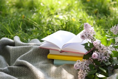 Photo of Stacked books, lilac flowers and blanket on green grass outdoors