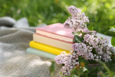 Photo of Stacked books, lilac flowers and blanket on green grass outdoors, closeup