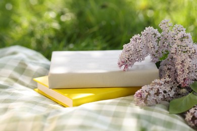Photo of Stacked books, lilac flowers and blanket on green grass outdoors, closeup