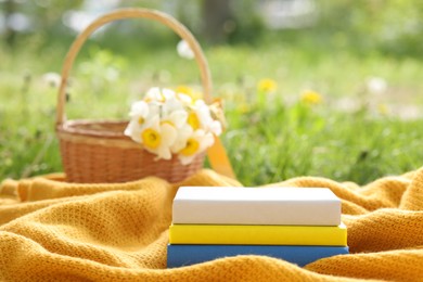 Photo of Stacked books, blanket and wicker basket with daffodil flowers on green grass outdoors, closeup
