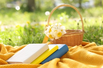 Photo of Stacked books, blanket and wicker basket with daffodil flowers on green grass outdoors, closeup