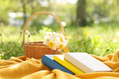 Photo of Stacked books, blanket and wicker basket with daffodil flowers on green grass outdoors, closeup