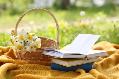 Photo of Stacked books, blanket and wicker basket with daffodil flowers on green grass outdoors