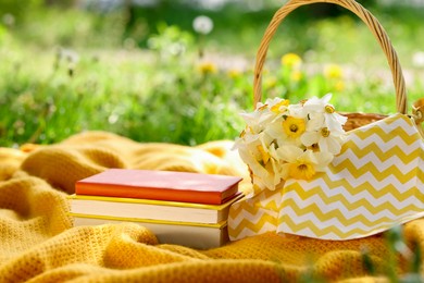 Photo of Stacked books, blanket and wicker basket with daffodil flowers on green grass outdoors, closeup