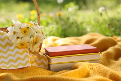 Photo of Stacked books, blanket and wicker basket with daffodil flowers on green grass outdoors, closeup
