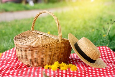 Photo of Wicker basket, dandelion flowers, straw hat and blanket on green grass outdoors