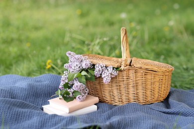 Photo of Stacked books, blanket and wicker basket with lilac flowers on green grass outdoors, closeup