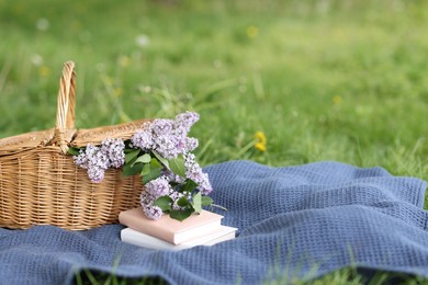Photo of Stacked books, blanket and wicker basket with lilac flowers on green grass outdoors, closeup. Space for text