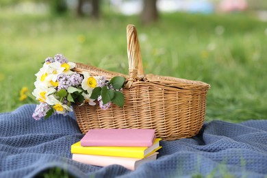 Photo of Stacked books, blanket and wicker basket with flowers on green grass outdoors, closeup