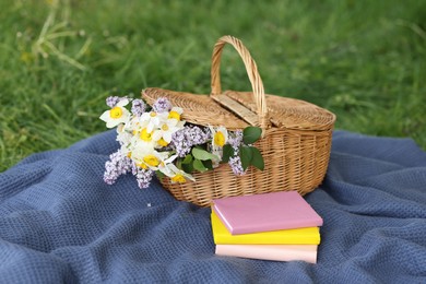 Photo of Stacked books, blanket and wicker basket with flowers on green grass outdoors