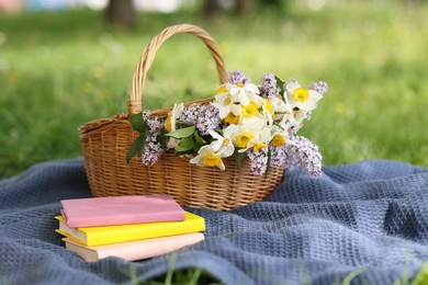 Photo of Stacked books, blanket and wicker basket with flowers on green grass outdoors
