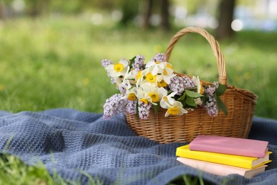 Photo of Stacked books, blanket and wicker basket with flowers on green grass outdoors