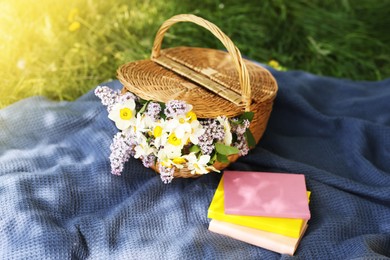 Photo of Stacked books, blanket and wicker basket with flowers on green grass outdoors