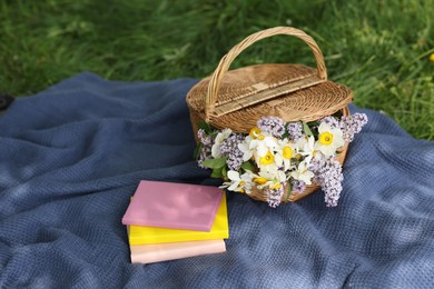 Photo of Stacked books, blanket and wicker basket with flowers on green grass outdoors