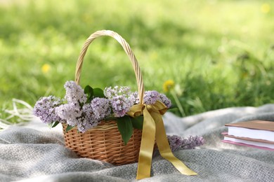 Photo of Stacked books and wicker basket with lilac flowers on green grass outdoors, closeup