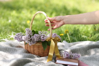 Photo of Woman with wicker basket, lilac flowers and books on blanket outdoors, closeup