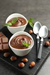 Photo of Tasty chocolate spread, pieces, mint and hazelnuts on gray table, closeup