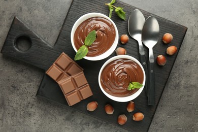Photo of Tasty chocolate spread, pieces, mint and hazelnuts on gray textured table, top view