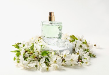 Photo of Bottle of aromatic perfume on podium and twigs with beautiful flowers against white background, closeup. Stylish presentation of product