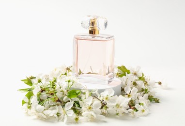 Photo of Bottle of aromatic perfume on podium and twigs with beautiful flowers against white background, closeup. Stylish presentation of product