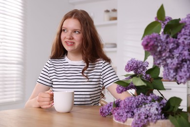 Photo of Smiling teenage girl with cup of hot drink and lilac flowers at table indoors