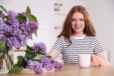 Photo of Smiling teenage girl with cup of hot drink and lilac flowers at table indoors