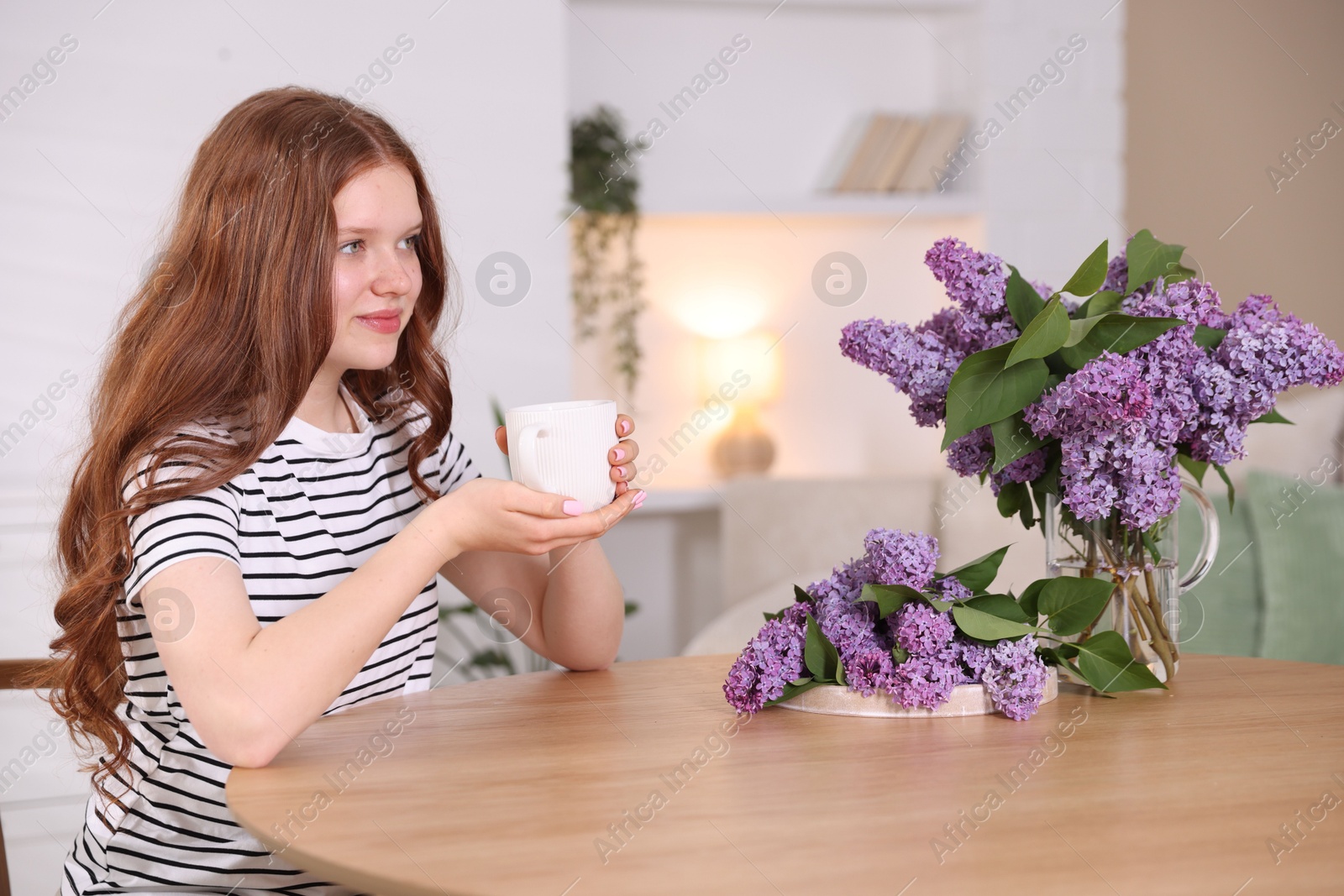 Beautiful teenage girl with cup of hot drink and lilac flowers at table indoors Photo of Beautiful teenage girl with cup of hot drink and lilac flowers at table indoors