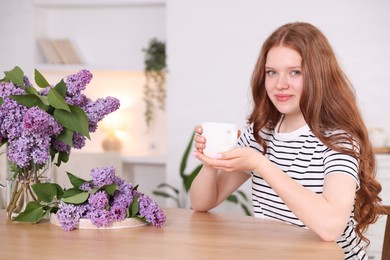 Photo of Beautiful teenage girl with cup of hot drink and lilac flowers at table indoors