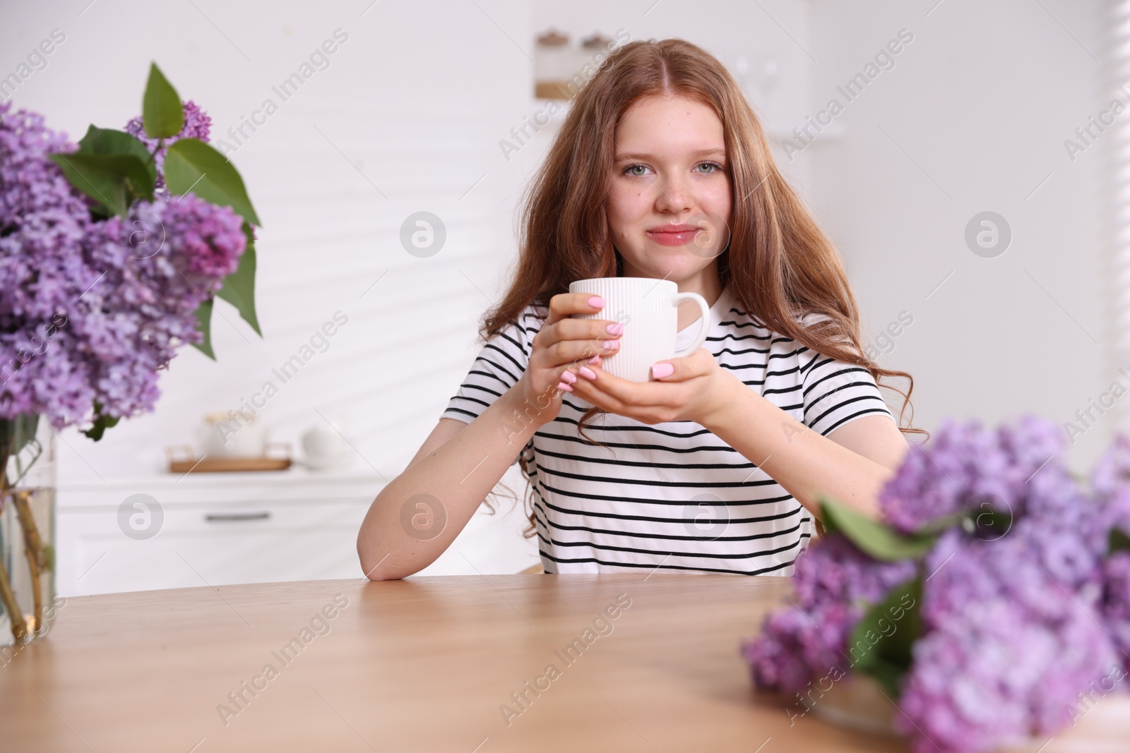 Beautiful teenage girl with cup of hot drink and lilac flowers at table indoors Photo of Beautiful teenage girl with cup of hot drink and lilac flowers at table indoors