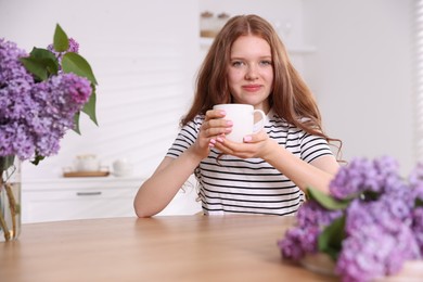 Photo of Beautiful teenage girl with cup of hot drink and lilac flowers at table indoors