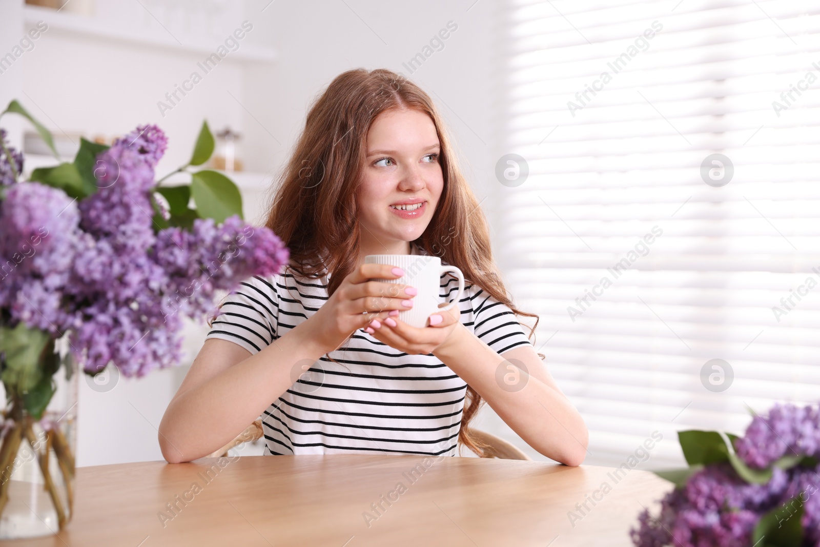 Smiling teenage girl with cup of hot drink and lilac flowers at table indoors Photo of Smiling teenage girl with cup of hot drink and lilac flowers at table indoors
