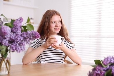 Photo of Smiling teenage girl with cup of hot drink and lilac flowers at table indoors