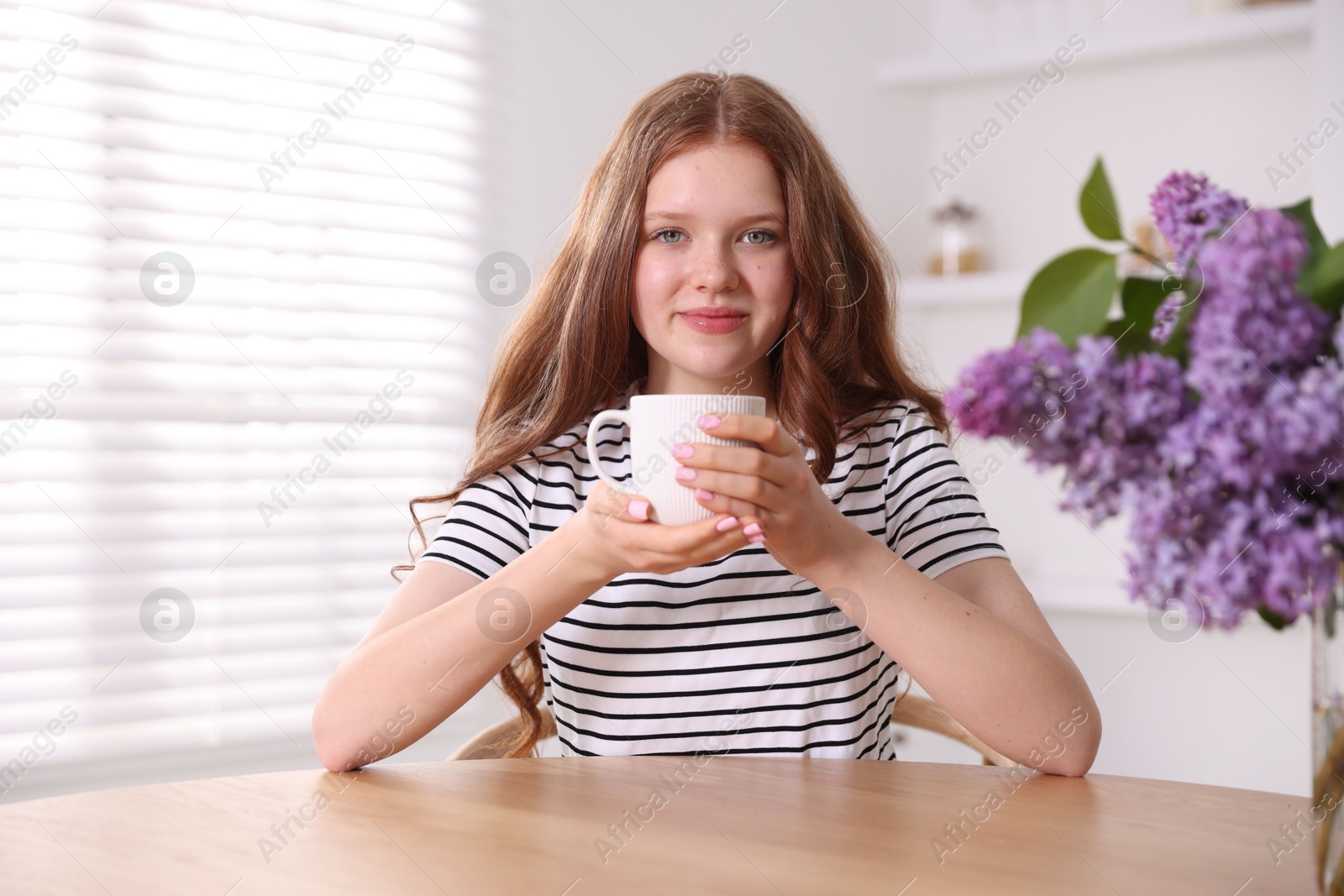 Beautiful teenage girl with cup of hot drink and lilac flowers at table indoors Photo of Beautiful teenage girl with cup of hot drink and lilac flowers at table indoors