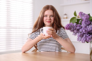 Photo of Beautiful teenage girl with cup of hot drink and lilac flowers at table indoors