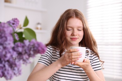 Photo of Beautiful teenage girl with cup of hot drink and lilac flowers at home