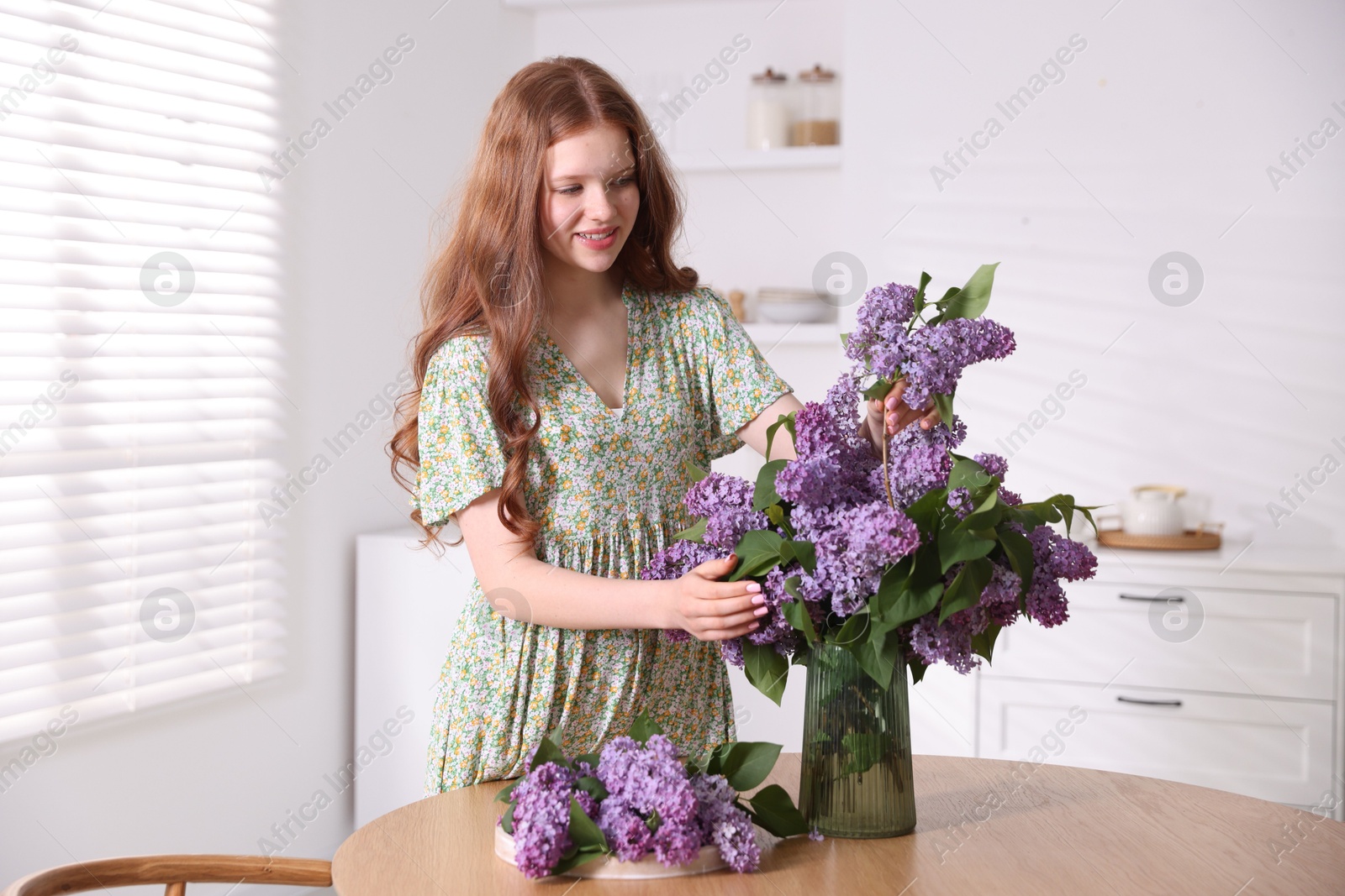 Smiling teenage girl putting branch with lilac flowers into vase at table indoors Photo of Smiling teenage girl putting branch with lilac flowers into vase at table indoors
