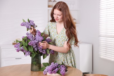 Photo of Beautiful teenage girl putting branch with lilac flowers into vase at table indoors