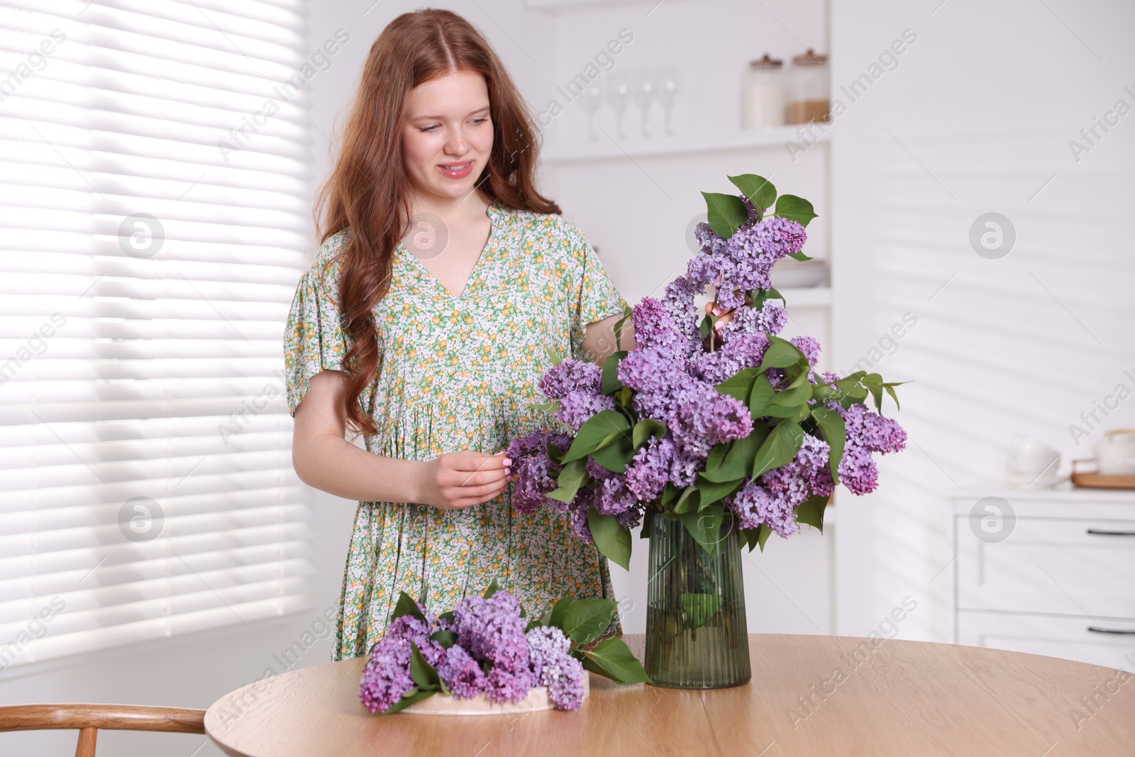 Smiling teenage girl putting branch with lilac flowers into vase at table indoors Photo of Smiling teenage girl putting branch with lilac flowers into vase at table indoors