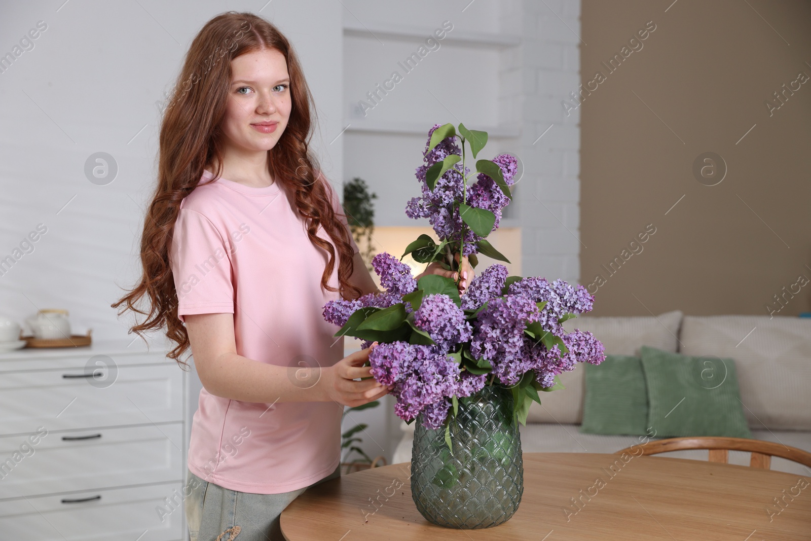 Beautiful teenage girl putting branch with lilac flowers into vase at table indoors Photo of Beautiful teenage girl putting branch with lilac flowers into vase at table indoors